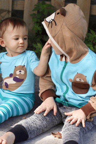 Two toddlers wearing matching otter-themed outfits, sitting outdoors and interacting.