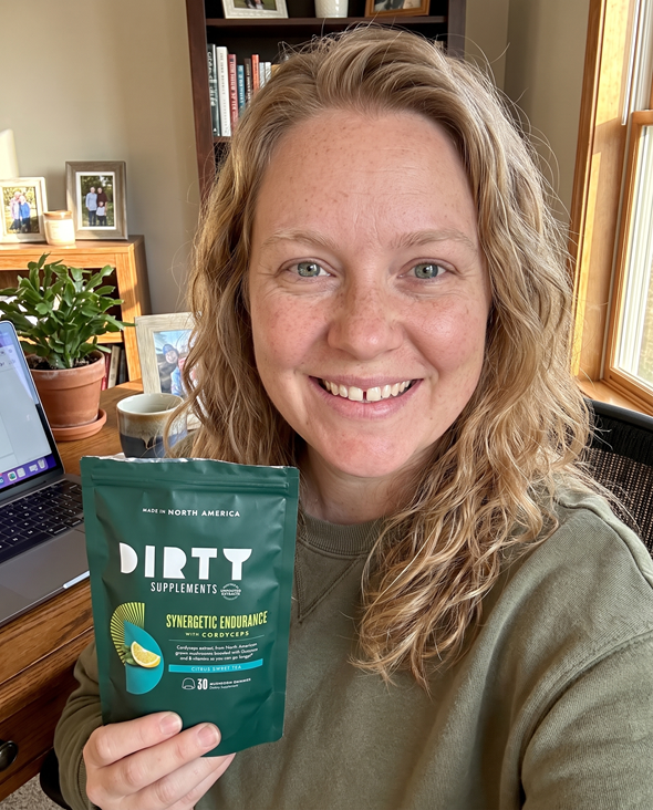 Woman in a kitchen holding DIRTY Supplements Synergetic Endurance citrus powder with cordyceps supplement packaging visible.